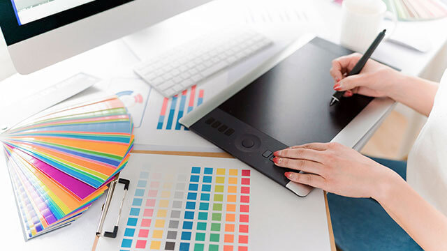 ペンタブレットでデザインする手と、PCの前のカラーチャート。私たちのクリエイティブな作業風景の一コマ。